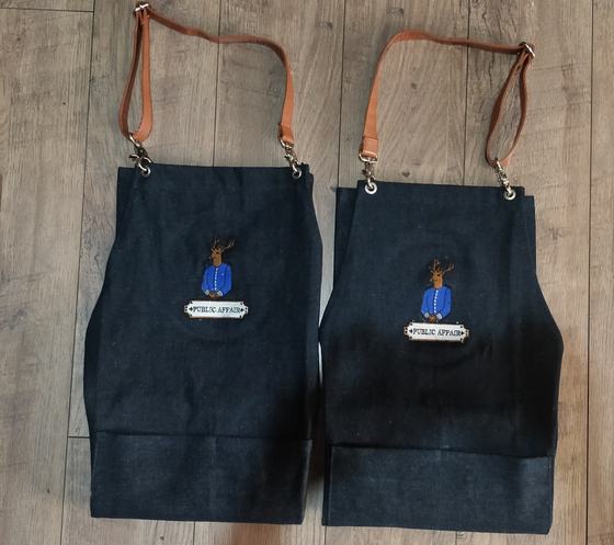Two dark denim aprons with tan leather straps and embroidered mascot badge logo, custom made for Public Affair by RestroCart, New Delhi