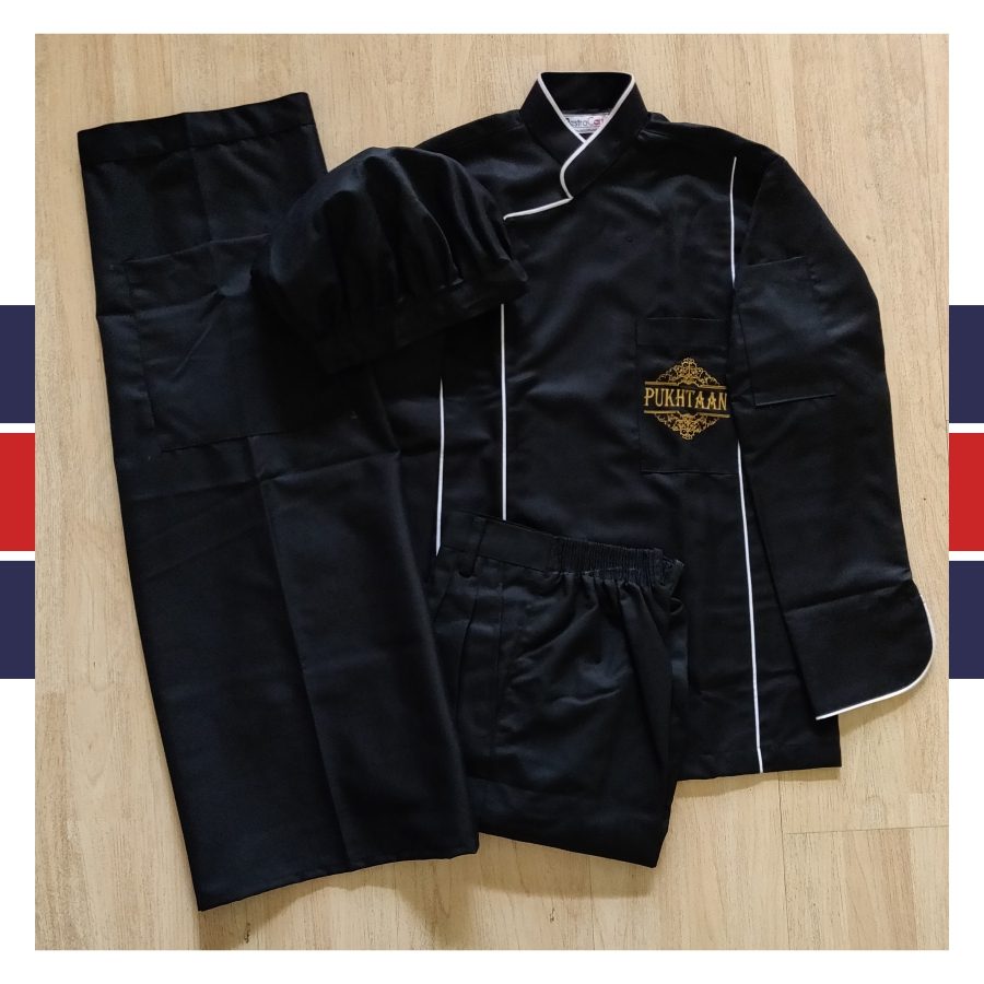 Black chef coat with white piping and a gold embroidered Pukhtaan logo, laid out with black trousers, chef uniform made for Pukhtaan by RestroCart
