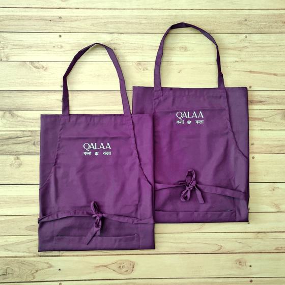 Two purple bib aprons with white embroidered logo and Hindi text, waist ties, laid flat, custom made for Qalaa by RestroCart, New Delhi