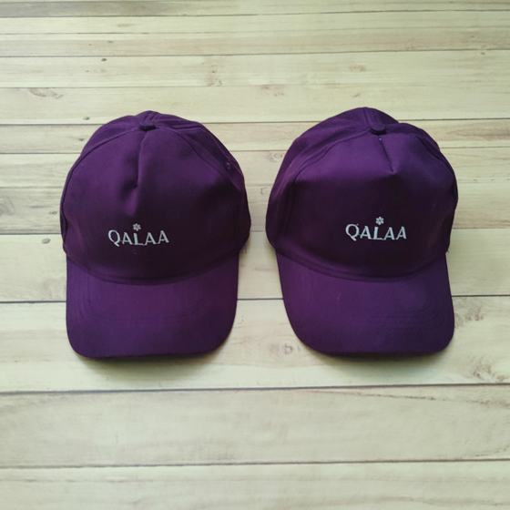 Two purple baseball caps with white embroidered logo on front, on light wooden floor, custom made for Qalaa by RestroCart, New Delhi