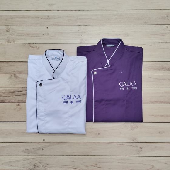 White and purple chef coats with contrast crossover piping and embroidered logo, folded side by side, custom made for Qalaa by RestroCart, New Delhi