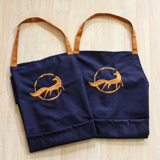 Two navy bib aprons with orange neck straps and large orange embroidered fox logo, laid flat, custom made for Quick Brown Fox by RestroCart, New Delhi