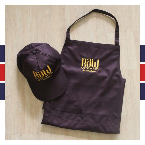 Dark brown bib apron and matching cap with gold embroidered logo, laid flat on light wood, custom made for Raw Cream by RestroCart, New Delhi