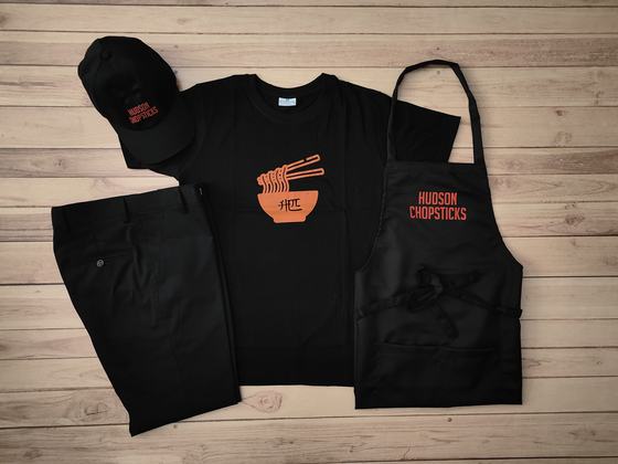 Black round neck t-shirt with orange noodle bowl print, black cap, trousers and bib apron, custom made by RestroCart, New Delhi