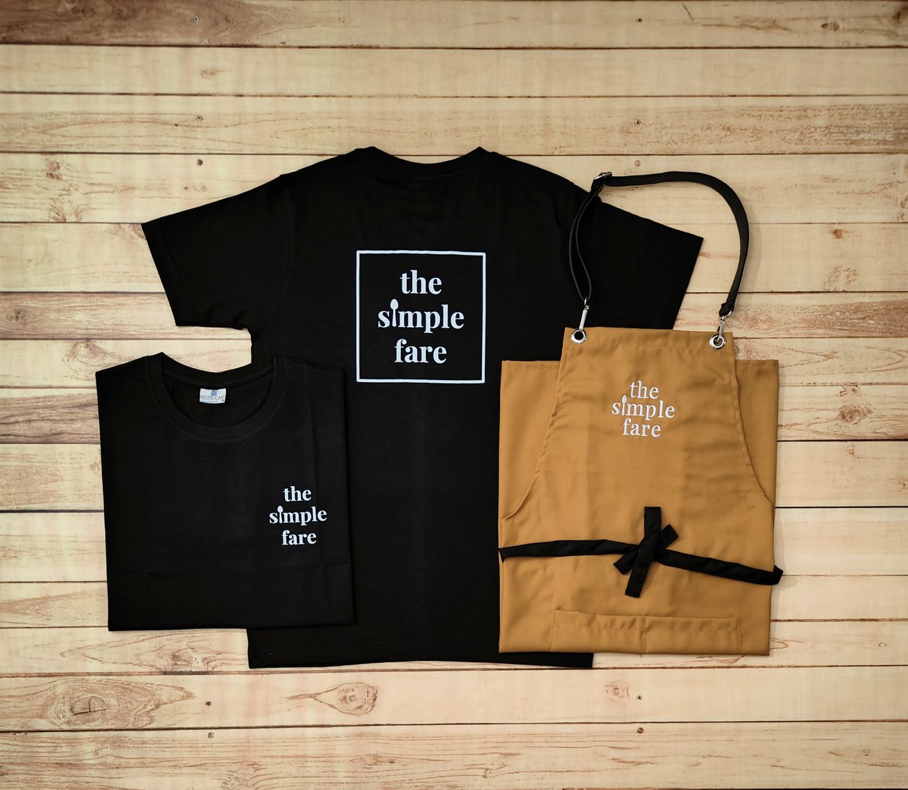 Black round-neck staff t-shirts with a large back print and small chest print, paired with a mustard bib apron carrying the same logo, cafe uniform set by RestroCart