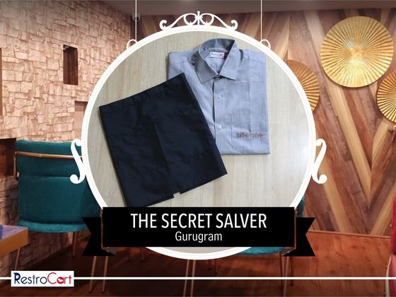 Black waist apron beside a folded grey long-sleeve service shirt with small logo, custom made for Secret Salver by RestroCart, New Delhi