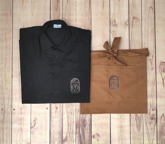 Black service shirt with embroidered arch logo on pocket beside a tan waist apron with matching logo, custom made by RestroCart, New Delhi