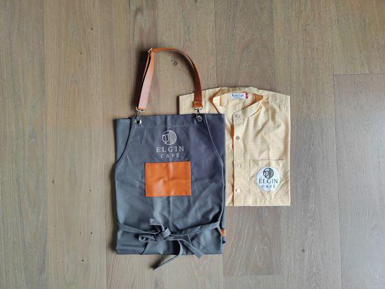 Grey bib apron with orange pocket and tan leather straps paired with pale yellow mandarin collar shirt, custom made for Elgin Cafe by RestroCart, New Delhi