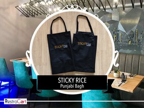 Two black bib aprons with gold printed logo, hanging side by side, custom made for Sticky Rice by RestroCart, New Delhi