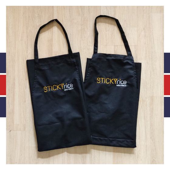 Two black bib aprons with orange and white embroidered logo on chest, laid flat on light wood, custom made for Sticky Rice by RestroCart, New Delhi