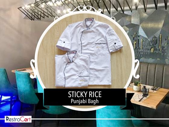 White short-sleeve chef coats with black piping and gold logo, one folded below, custom made for Sticky Rice by RestroCart, New Delhi