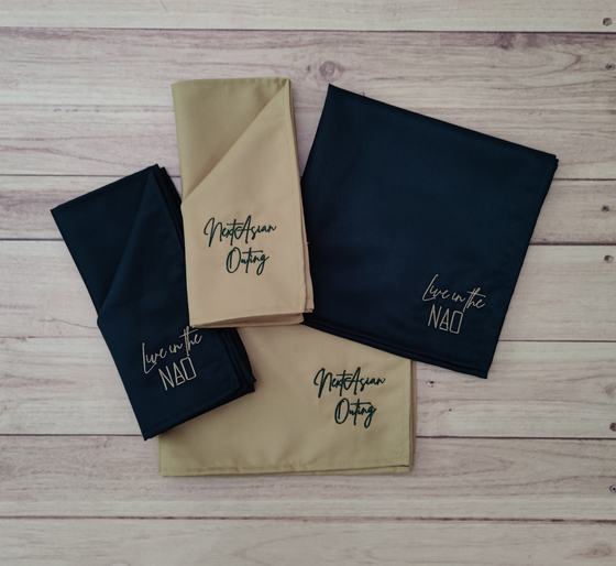 Navy and beige cloth table napkins with embroidered script slogans, folded and fanned out on wood, custom made by RestroCart, New Delhi
