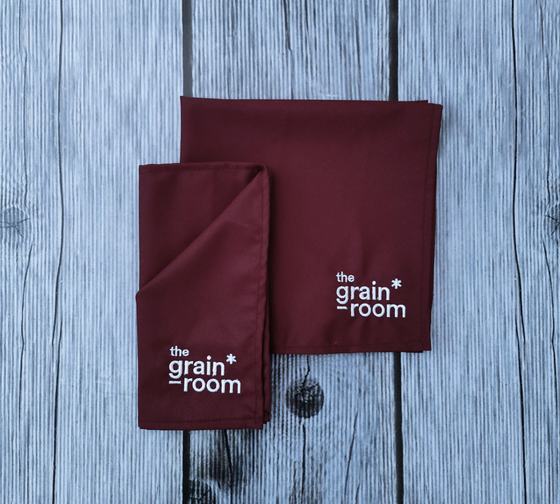 Pair of maroon cloth table napkins with white embroidered wordmark, one folded, on grey wood, custom made by RestroCart, New Delhi