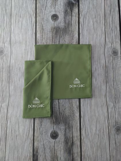 Pair of olive green cloth table napkins with white embroidered dome logo, one folded, on grey wood, custom made by RestroCart, New Delhi