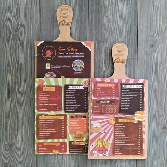 Colourful printed menu cards clipped to wooden paddle boards with engraved logo, laid on grey wood, custom made for Tapri by RestroCart, New Delhi