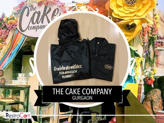 Black hooded raincoat with white hashtag and phone number print and a folded jacket, custom made for The Cake Company by RestroCart, New Delhi