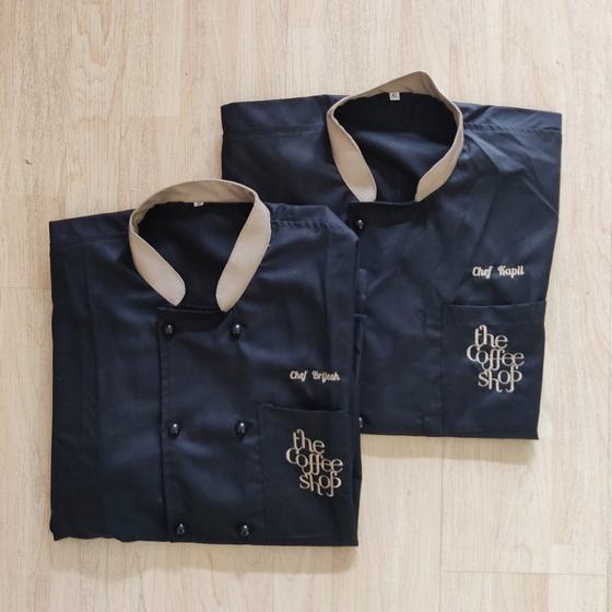 Two black chef coats with beige collar, gold embroidered chef names and script logo, folded, custom made for The Coffee Shop by RestroCart, New Delhi