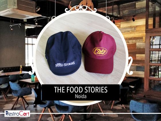Navy baseball cap with white printed logo beside a maroon cap with yellow logo, custom made for The Food Stories by RestroCart, New Delhi