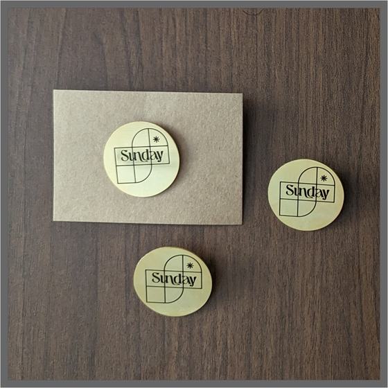 Three round brushed gold metal badges with black engraved logo, on kraft card and dark wood, custom made for The Sunday House by RestroCart, New Delhi