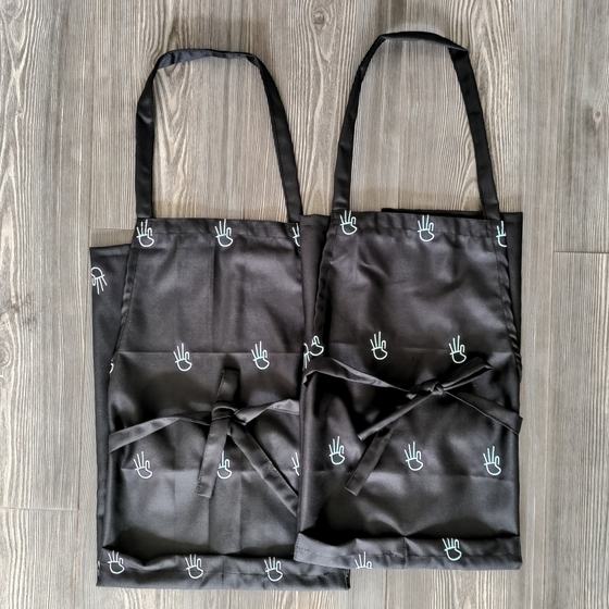 Two black satin bib aprons with all over white hand sign print and waist ties, laid flat, custom made for Third Roast by RestroCart, New Delhi