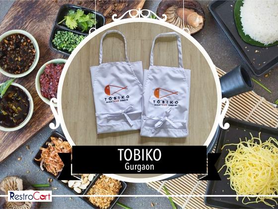 Two light grey bib aprons with orange and black printed logo and white ties, laid flat, custom made for Tobiko by RestroCart, New Delhi