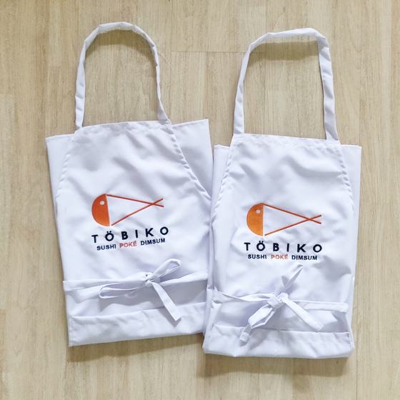 Two white bib aprons with orange and black printed fish logo and white waist ties, laid flat, custom made for Tobiko by RestroCart, New Delhi
