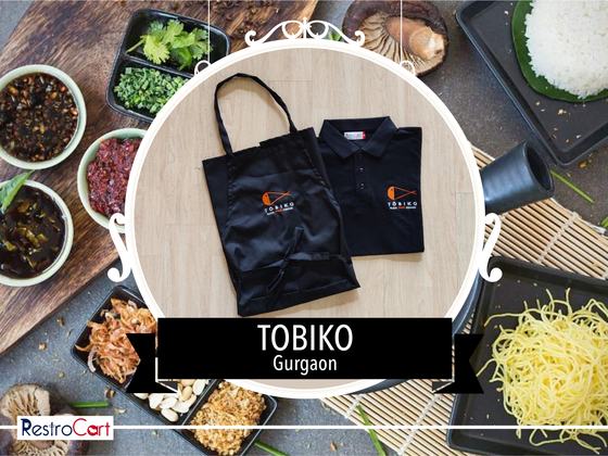Black bib apron with white logo beside a black polo t-shirt with small chest logo, custom made for Tobiko by RestroCart, New Delhi