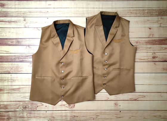 Pair of tan waistcoats with shawl lapels, gold embroidered logo and welt pockets, laid flat on wood, custom made by RestroCart, New Delhi