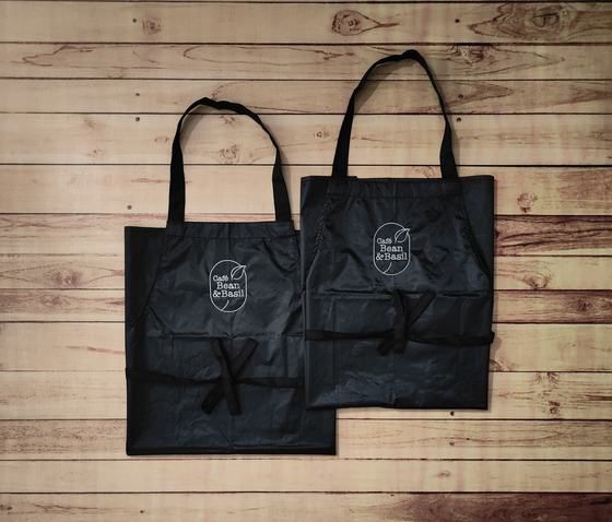 Pair of black waterproof bib aprons with white printed cafe logo and front pockets, laid flat on wood, custom made by RestroCart, New Delhi