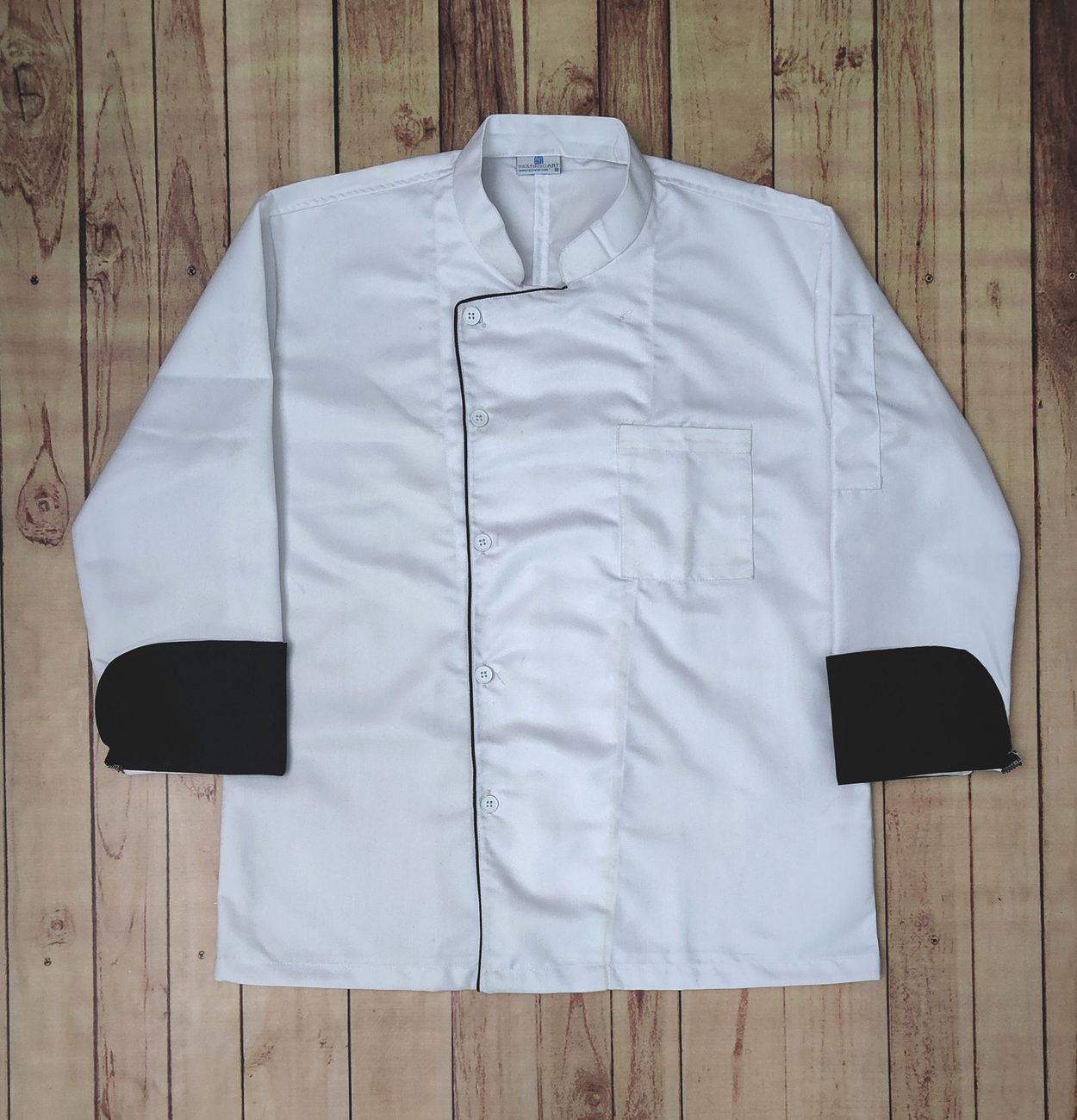 White full-sleeve chef coat with an offset front, thin black piping and black turn-back cuffs, laid flat on wooden boards