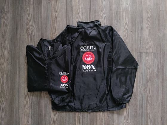 Black windcheater jackets with red round logo and white printed text, two laid flat, custom made for Eden by RestroCart, New Delhi