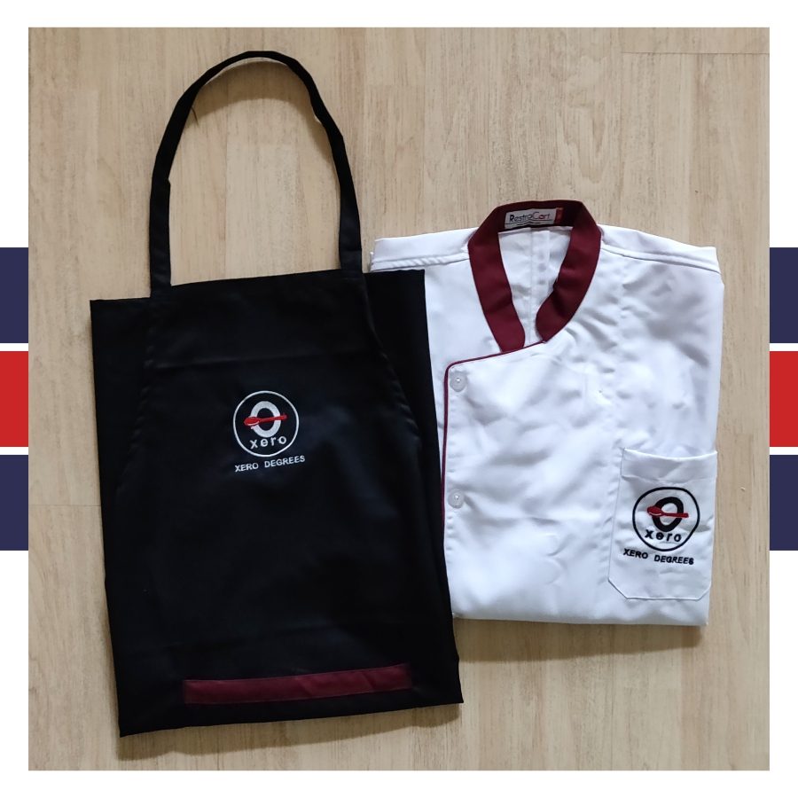 White chef coat with maroon collar and piping beside a black bib apron with a maroon hem band, both embroidered with the Xero Degrees logo